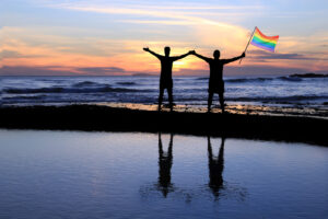 Silhouette of a gay couple holding a rainbow pride flag at sunset.